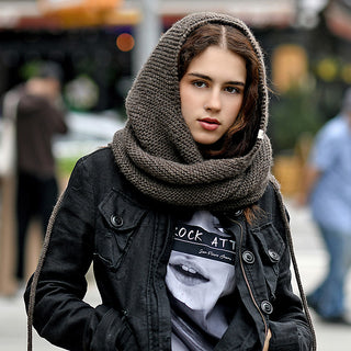 A young woman with long brown hair wears the Anna Hood—a thick, knitted brown scarf—over her head and neck, paired with a black jacket and printed t-shirt. She stands outdoors; people and street elements appear blurred in the background.