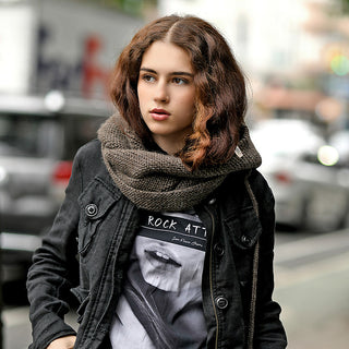 A young person with wavy brown hair stands outdoors in the Anna Hood, a dark jacket, chunky knit scarf, and graphic tee. The blurred city street background features cars and buildings.
