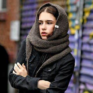 A person with light skin and wavy brown hair wears the Anna Hood chunky knit hooded scarf and a black jacket, standing with arms crossed in front of a graffiti-covered wall.