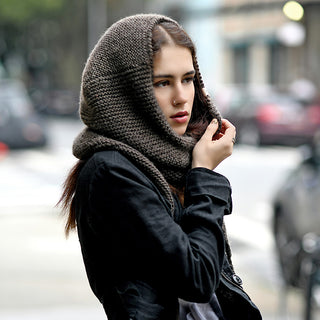 Wearing the Anna Hood, a person in a black coat stands outdoors on a city street with blurred parked cars and buildings in the background, looking to the side.