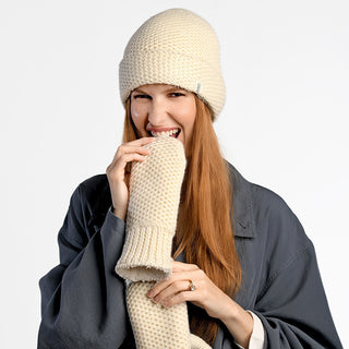 A woman with long red hair wears a cream knit hat and gray jacket, holding a handmade Nepalese scarf near her mouth, while cozy Kent Mittens peek out against a plain white background.