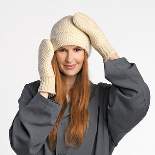 A woman with long red hair, wearing a gray coat, a cream knitted hat, and Kent Mittens, stands against a white background, smiling and touching her hat.