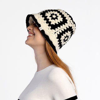 A person with long reddish hair smiles in profile against a white background while wearing the Milie Hat—a black and white, geometric-patterned, crocheted bucket hat handmade in Nepal.