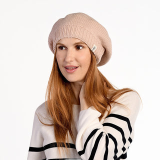 A woman with long red hair wears the Abbey Hat, a light pink wool beanie handmade in Nepal, along with a white sweater with black stripes. She stands against a plain white background, looking to the side and touching her neck.