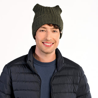 A person with light skin and brown, wavy hair smiles at the camera against a white background, wearing the Ely Hat—a dark green wool beanie—paired with a navy quilted jacket and a blue crewneck shirt.