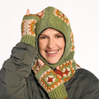 A person smiles at the camera wearing the Ryan Hood, a green, orange, and beige crochet balaclava handmade in Nepal, with matching fingerless gloves and a dark quilted jacket against a plain white background.