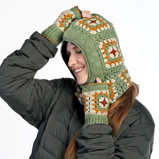 A person wearing the Ryan Hood, a handmade in Nepal crocheted green hood with multicolored granny square patterns, plus matching fingerless gloves and a dark puffer jacket, smiles and touches the top of the hood.