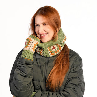 A woman with long red hair wears the Ryan Hood, a dark green puffer jacket, and a matching handmade multicolored crocheted scarf and fingerless gloves from Nepal, smiling with her hand on her face against a plain white background.