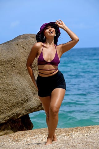 A woman wearing a purple sunhat, Bikini Top - Solid, and black shorts stands on a sandy beach near a large rock with the ocean and blue sky in the background.