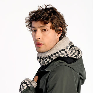 A person with curly brown hair wears the Ryan Hood—a dark green jacket with a wool hood—paired with a black and white checkered knitted scarf and matching gloves, looking over their shoulder against a plain background.