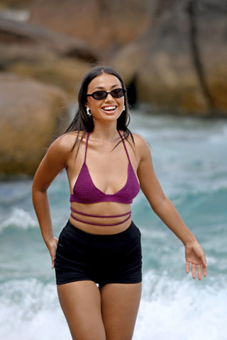 A woman wearing sunglasses, black shorts, and the Bikini Top - Solid—a hand-crocheted purple bikini with a classic triangle design—stands in shallow beach water, smiling with waves and rocks in the background.