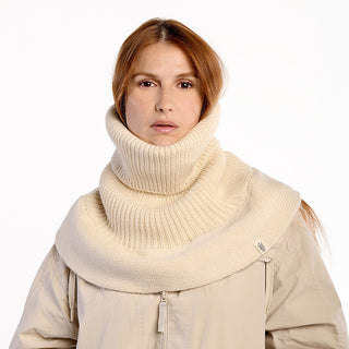 A person with long auburn hair wears the Hank Neckwarmer, a thick cream-colored ribbed knit cowl over a beige coat, covering the neck and shoulders. The background is plain white.