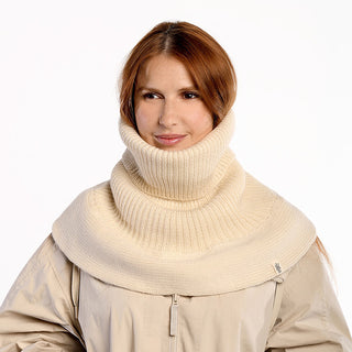 A person with long, reddish-brown hair wears the Hank Neckwarmer—a beige ribbed knit scarf—over a light beige jacket, standing in front of a plain white background.