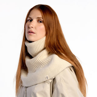 A woman with long straight red hair wears the Hank Neckwarmer and a matching cream-colored coat, looking slightly to the side against a plain white background.