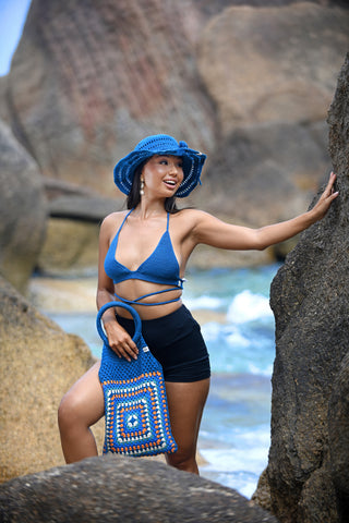 A woman stands outdoors among large rocks by the ocean, smiling and looking to the side. She wears a blue sun hat, blue bikini top, black shorts, and holds a Cotton Crochet Handbag in blue and orange.