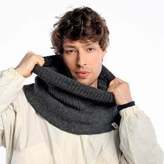 A person with wavy brown hair wears a light jacket and pulls up a thick, dark gray Hank Neckwarmer around their neck, looking directly at the camera against a plain white background.