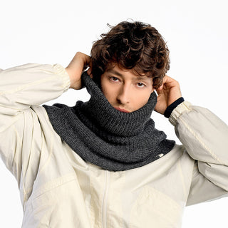 A person wearing a light-colored jacket pulls up the Hank Neckwarmer, a thick dark gray knit accessory, over their mouth and neck against a plain white background.