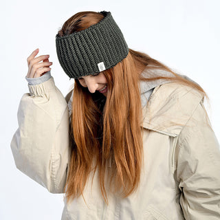 A person with long red hair wears a thick beige winter coat and the Mandy Headband, adjusting it with one hand. The person's head is tilted down against a plain white background.