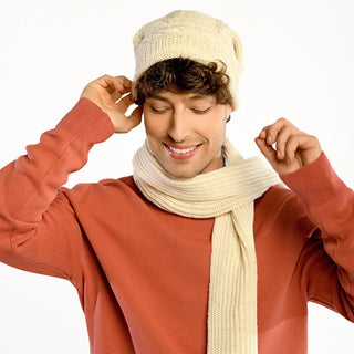 Wearing the Gateway Hat—handmade in Nepal from 100% wool in cream—and a matching scarf with a salmon sweater, a smiling person stands against a plain white background, looking downward.