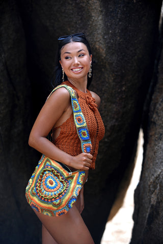 A woman stands outdoors between large rocks, smiling with sunglasses on her head, wearing a sleeveless brown crochet top, shorts, and carrying the Cotton Crochet Messenger Bag on her shoulder.