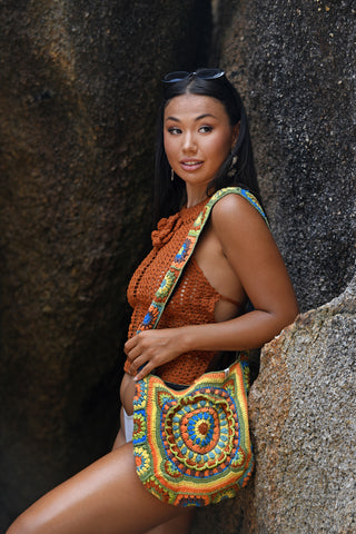 A woman stands outdoors by large rocks, wearing a rust crochet top, white bottoms, and carrying the Cotton Crochet Messenger Bag. Sunglasses rest on her head as she gazes slightly to the side.