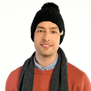 A man wearing the Cedar Hat, a handmade black wool beanie with a pom-pom from Nepal, paired with a dark gray scarf, light blue shirt, and rust sweater, stands against a plain white background.