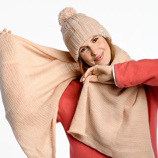 A woman in a pink sweater smiles while spreading out the Air Wrap Scarf, paired with a beige knit pom-pom hat. She poses in front of a plain white background.