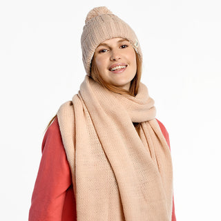 A woman smiles at the camera, wearing an Air Wrap Scarf and matching hat in light beige merino wool over a pink sweatshirt, standing against a plain white background.