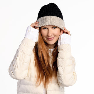 A woman with long reddish-brown hair wears the handmade Cliff Hat, a black and beige wool beanie from Nepal, along with a puffy off-white jacket. She smiles softly, adjusting her hat with both hands against a plain white background.
