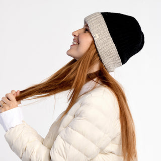 A person wearing the Cliff Hat, a black and beige wool beanie, and a white puffer jacket smiles while holding their long, straight hair in profile against a plain white background.
