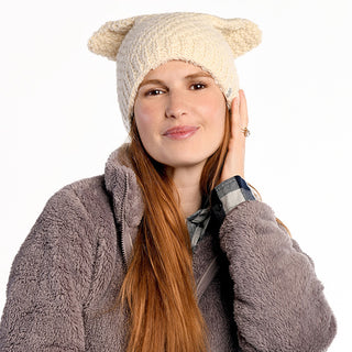 A woman with long red hair wears a cream Bear Ear Beanie wool winter hat and a gray fleece jacket over a plaid shirt, touching her hat and smiling gently against a plain white background.