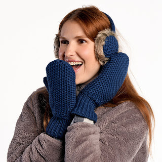 Wearing Linda Earmuffs, a person in a brown fuzzy coat and dark blue knit mittens holds their hands to their face and smiles against a plain white background.