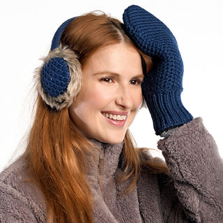 A woman with long red hair wears Linda Earmuffs, a fuzzy gray coat, and blue knit mittens, smiling as she holds one mittened hand to her forehead against a white background.