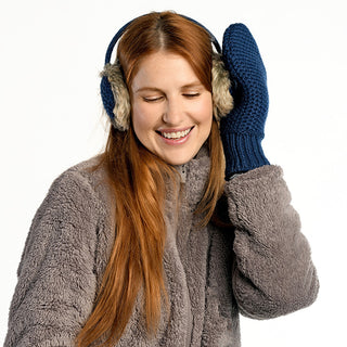 A woman with long red hair wears tan Linda Earmuffs, a gray fuzzy jacket, and blue knit mittens. She smiles with her eyes closed, holding one mitten to her Linda Earmuffs. The background is white.