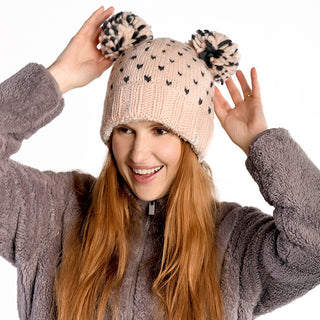 A person with long red hair smiles while wearing the Double Pom Kira Hat, a beige wool hat with black dots and two large pom-poms, paired with a fuzzy grey sherpa-lined jacket against a plain white background.