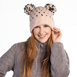 A woman with long red hair smiles and touches the brim of her Double Pom Kira Hat—light pink wool with sherpa lining, two large pom-poms, and black dots—paired with a gray zip-up fleece jacket. The hat is handmade in Nepal.