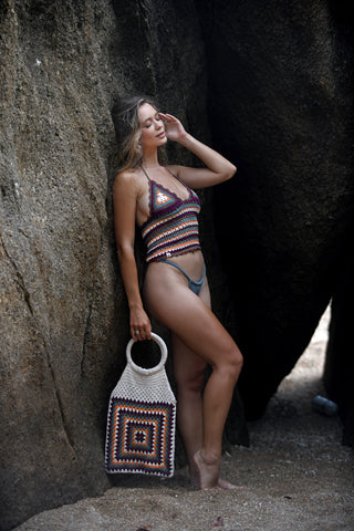A woman in a multicolored Striped Classic Top stands barefoot between large rocks on a sandy beach, holding a matching crochet bag and touching her hair while gazing into the distance.