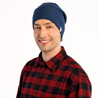 A person is smiling while wearing The Depp Slouch, a blue slouchy beanie, and a red and black plaid shirt against a plain white background.