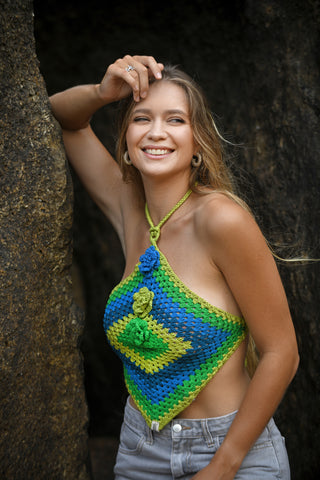 A woman with long hair smiles between two rocks, wearing the Diamond Top—a halter-style crocheted top with geometric blue, green, and yellow patterns—paired with light gray jeans. She raises one hand to rest on her head.
