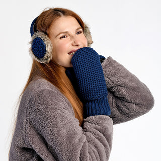 A woman with long red hair smiles while holding a blue knit mitten close to her face, wearing Linda Earmuffs and a gray fuzzy coat against a plain white background.