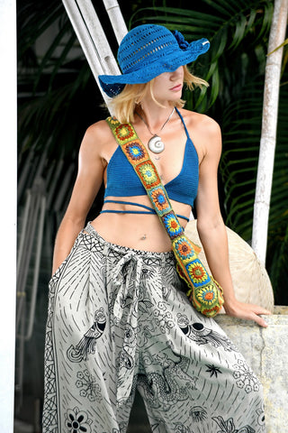 A person stands outdoors with one hand on a wall, in front of leafy greenery, wearing a blue sun hat, blue crochet halter top, patterned wide-leg pants, and carrying the Cotton Crochet Messenger Bag.