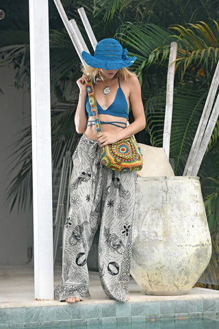 By the pool, a person holds a Cotton Crochet Messenger Bag while wearing a blue sunhat, blue bikini top, and wide-leg printed pants. Large potted plants and lush greenery fill the background.