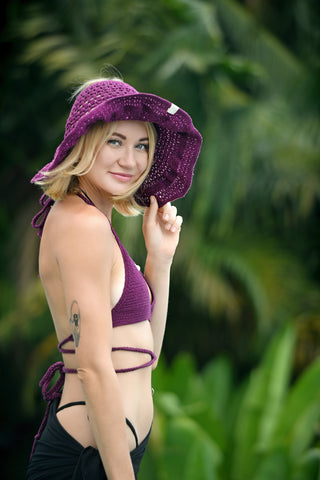 A woman wearing an Open Top Sun Hat and a purple bikini top stands outdoors, looking at the camera with tropical greenery in the background.