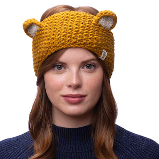 A person with auburn hair is wearing the Ears Headband, a yellow, knitted accessory featuring small, protruding bear ears. This charming headband, handmade in Nepal from 100% wool, complements their dark blue sweater. The person has a neutral expression, looking directly at the camera against a plain white background.