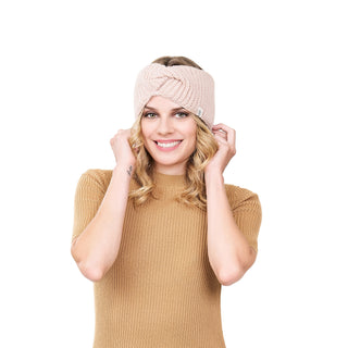 A person with long blonde hair is smiling and looking forward while wearing a pink Tuk Headband made of hand-knit merino wool and a brown short-sleeve knit top. The fashionable design stands out against the plain white background.