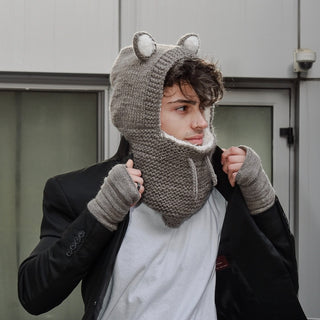 A person with tousled hair is wearing a gray knitted hat made of Merino wool with bear ears and a matching scarf that covers the lower part of their face. They are adjusting their hat with both hands, revealing Reverse Step Handwarmers, dressed in a white shirt and dark jacket. Behind them is a building with a glass door.