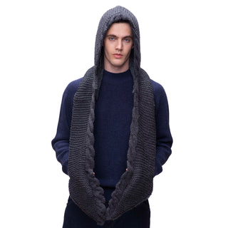 A man with a serious expression is wearing a dark blue sweater and a chunky knit 2 Pocket Scarf Hood in grey, which also serves as a hood. Handmade in Nepal from merino wool, this versatile scarf drapes down over his shoulders and chest against a plain white background.