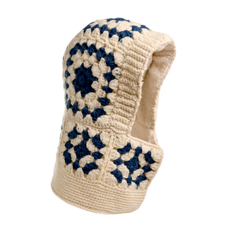 The Crochet Patch Hood is a handmade cream-colored hood adorned with blue geometric crochet patterns, and it features a lined interior for added warmth. It's designed to cover both the head and neck.