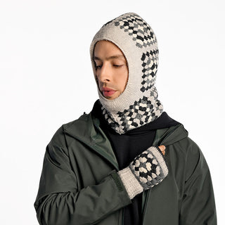Wearing a green jacket, black shirt, and the handmade-in-Nepal Ryan Hood—a beige-and-black checkered knitted wool balaclava with matching fingerless gloves—a person looks down against a plain white background.