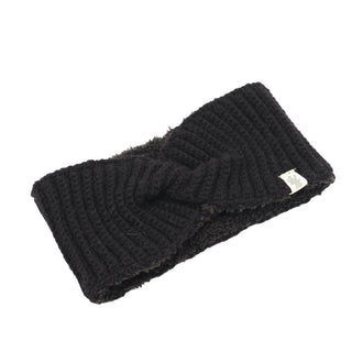 The Tuk Headband is a black, hand-knit accessory with a trendy twisted design at the center. Crafted from soft merino wool, it features a small white tag on one side and has a plush inner surface for added comfort. It is showcased on a plain white background.